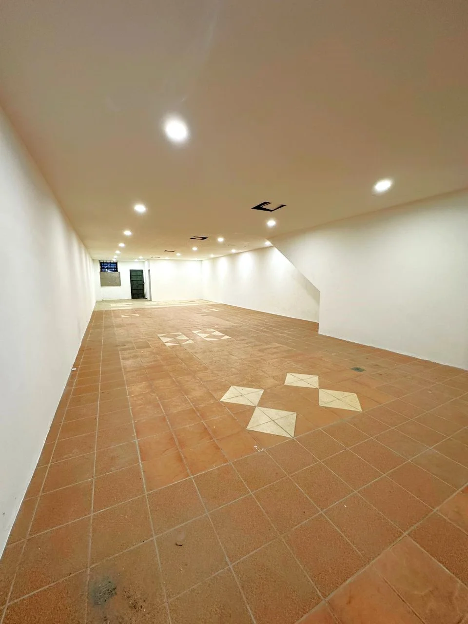 Room features: white painted walls, tile flooring with decorative pattern, recessed LED downlights, open layout, no visible windows, smooth ceiling, corner wall with slight angle; style: modern.